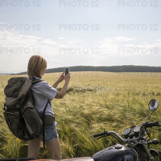 Caucasian woman standing in field near motorcycle using cell phone