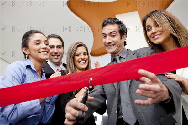 Hispanic co-workers watching businessman cutting red ribbon