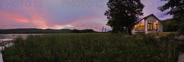 Panoramic shot of an illuminated cabin with clapboard siding by lake at dusk