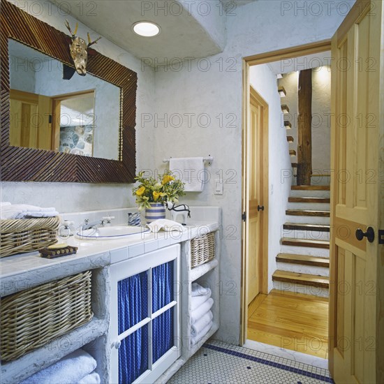 View of rustic wood stairway through open door on right. White open cabinets on left with wicker baskets holding white cotton greek  key pattern towels. White ceramic tile and yellow calla lilies. Rustic framed mirror with animal skull.