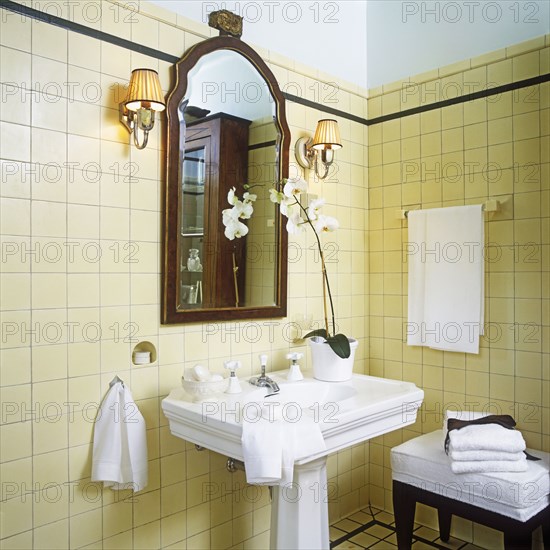 1920s style. Detail of white stately pedestal sink in center and mahogany framed mirror. White orchids sitting on right side of sink. Shade sconces. Cream