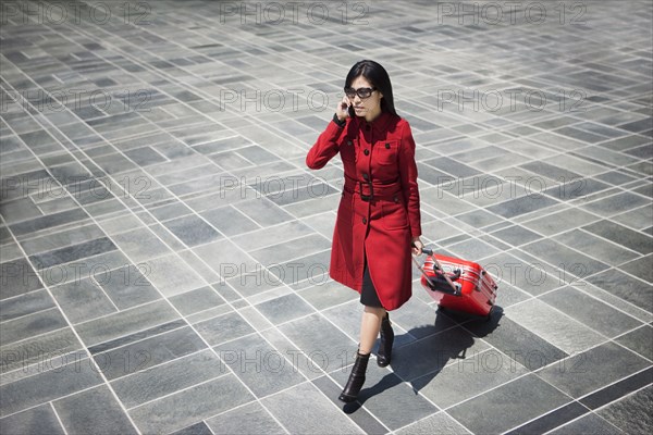 Asian woman pulling rolling suitcase and talking on cell phone