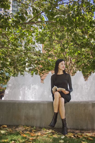 Asian woman sitting outdoors on concrete wall reading book