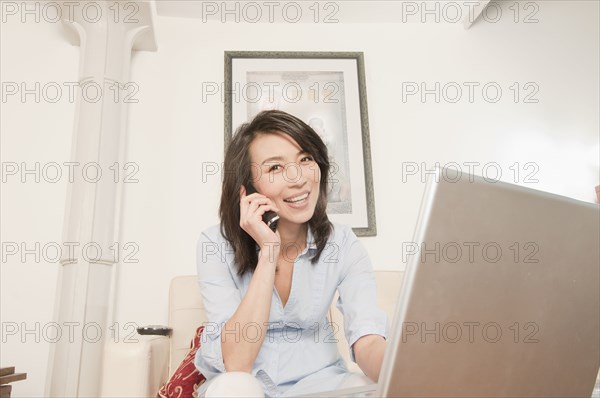 Korean woman using laptop and talking on phone in living room