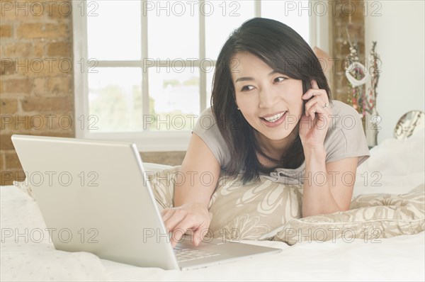 Korean woman laying on bed using laptop and talking on phone