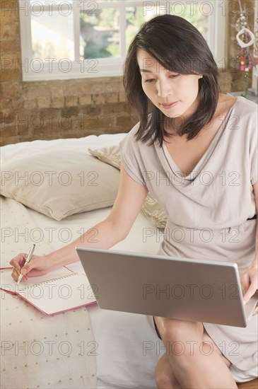 Korean woman sitting on bed using laptop and writing