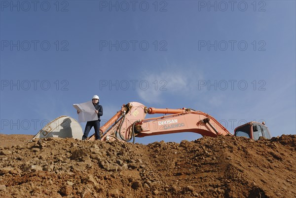 Female engineer holding blueprint standing in front of excavator low angle view