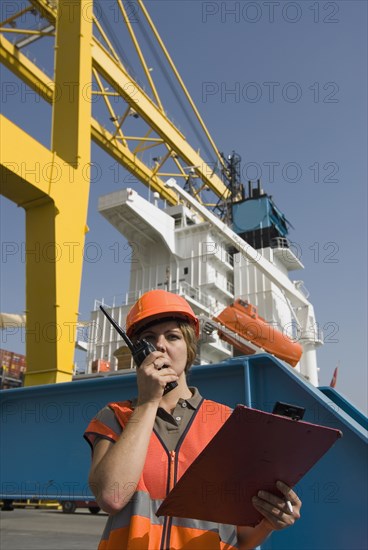 Woman wearing hard hat standing by container ship using walkie-talkie