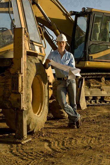 Young engineer with blueprint by excavator at construction site