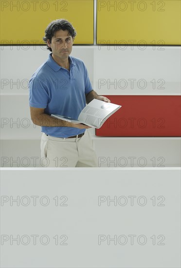 Male office worker holding book in front of coloured shelves portrait