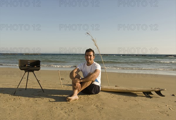 man sitting on beach with surfboard and barbecue