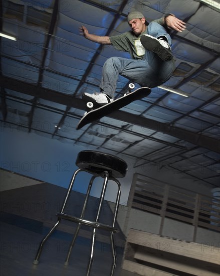 Caucasian man jumping on skateboard over stool indoors