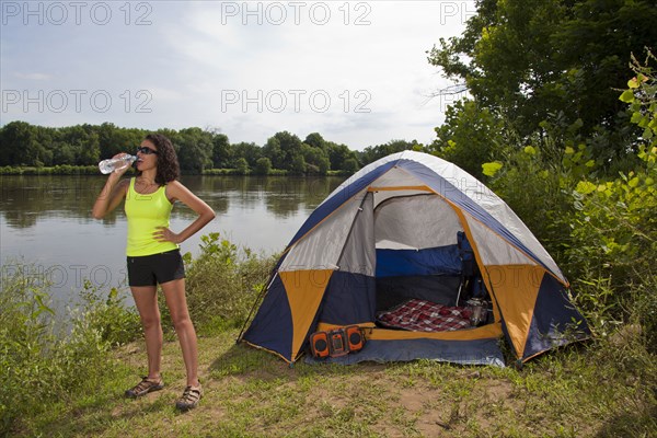 Hispanic woman drinking water near camping tent at lake