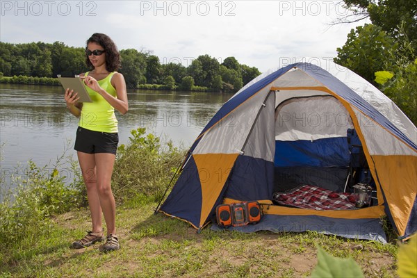 Hispanic woman using digital tablet near camping tent at lake