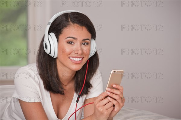 Hispanic woman laying on bed listening to cell phone with headphones