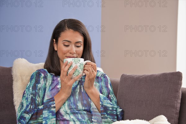 Hispanic woman sitting on sofa drinking warm beverage