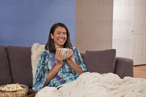 Laughing Hispanic woman sitting on sofa drinking warm beverage