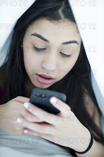 Hispanic girl using cell phone under blanket