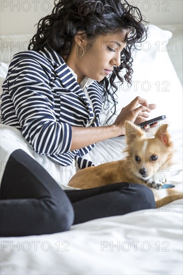 Mixed race woman using cell phone with dog on bed