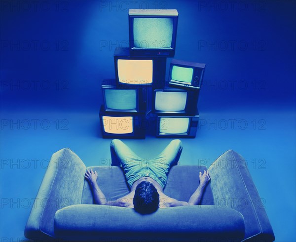 Man sitting on loveseat watching stack of televisions