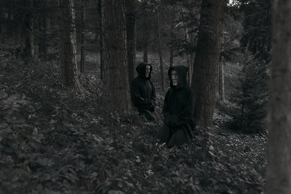 People wearing black robes and white masks sitting in forest
