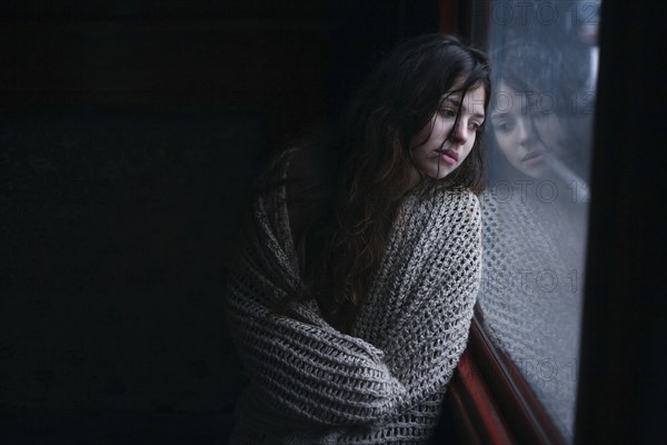 Sad Caucasian woman wrapped in scarf leaning on window