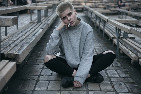 Punk Caucasian woman sitting on cobblestones smoking cigarette