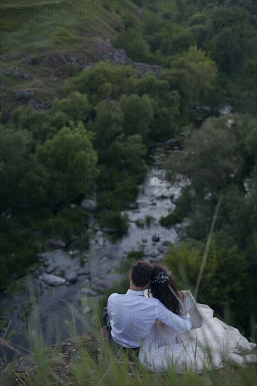 Caucasian bride and groom sitting on hill admiring river