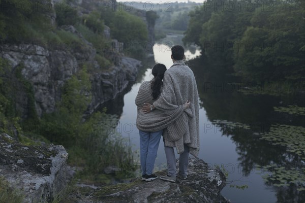 Caucasian couple wrapped in blanket standing on rock admiring river