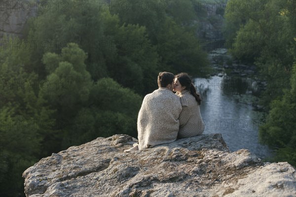 Caucasian couple wrapped in blanket kissing on rock near river