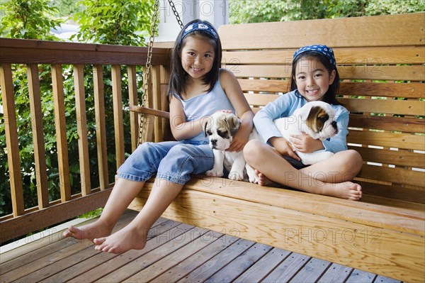 Young Asian sisters sitting on porch swing with puppies