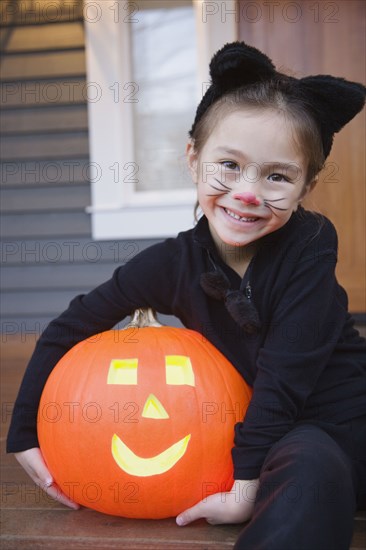 Mixed race young girl in cat costume holding Halloween pumpkin