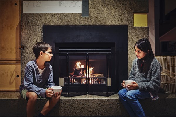 Mixed race children having hot drink together near fireplace