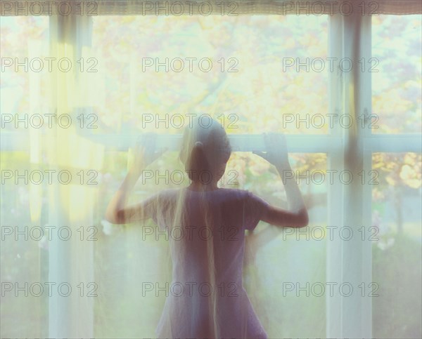 Mixed race girl standing behind sheer curtain in window