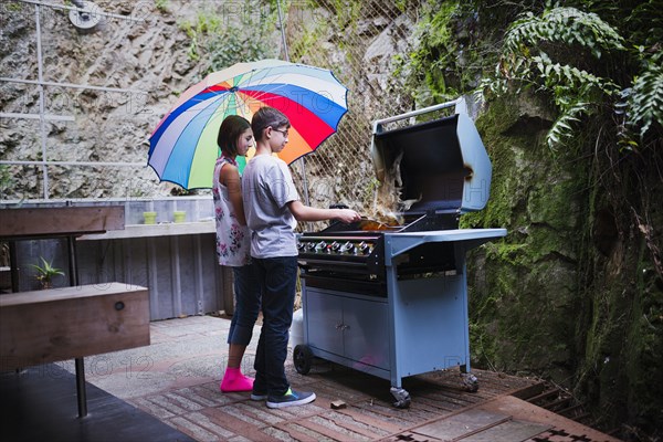 Mixed Race brother and sister cooking on patio grille under umbrella