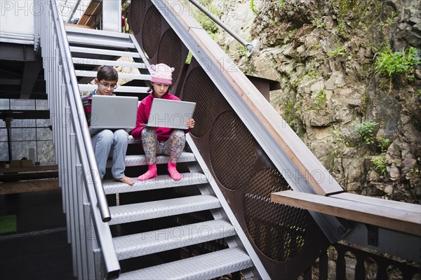 Mixed Race brother and sister sitting on staircase using laptops