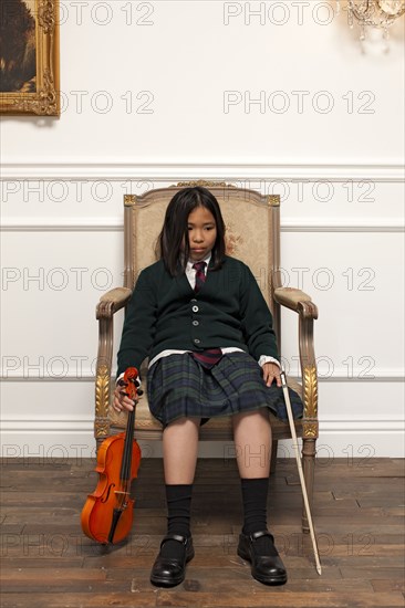 Sad Vietnamese girl sitting in elegant chair holding violin