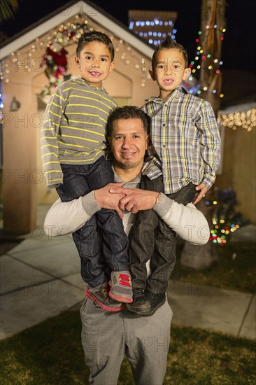 Hispanic man carrying sons on shoulders outside house decorated with string lights