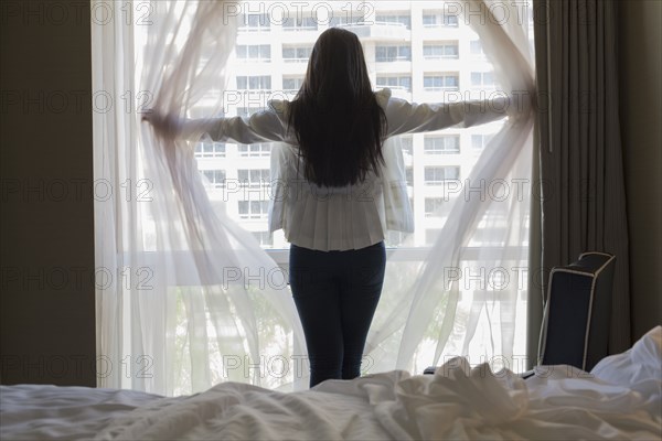 Asian teenage girl opening curtains at hotel window