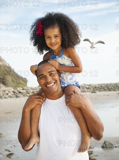 Black father carrying daughter on shoulders on beach