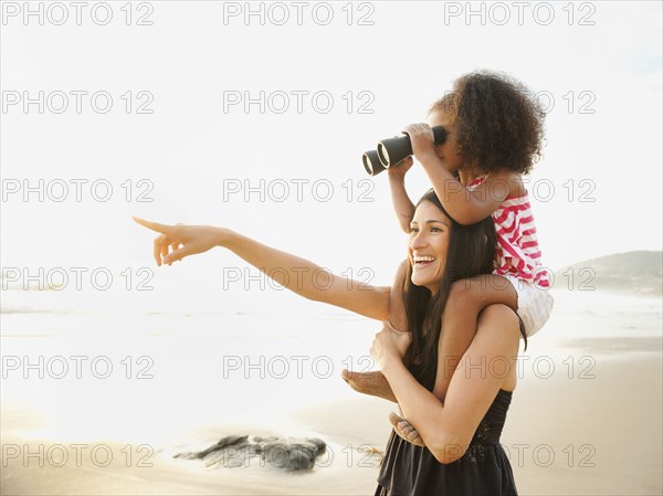 Hispanic mother carrying daughter with binoculars on beach