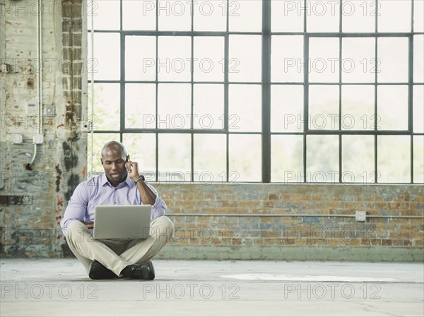 Black businessman using laptop and cell phone on warehouse floor