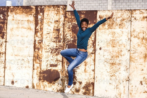 African American woman jumping for joy near rusty metal wall