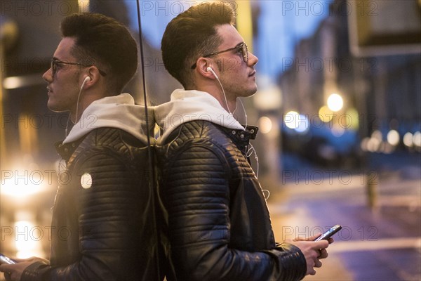 Pensive Caucasian man leaning on wall listening to music on cell phone