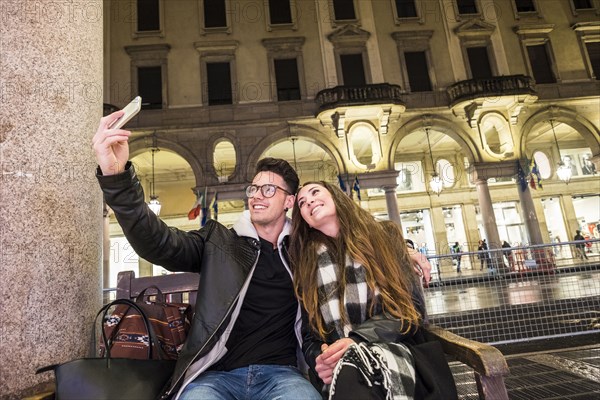 Caucasian couple sitting on urban bench posing for cell phone selfie