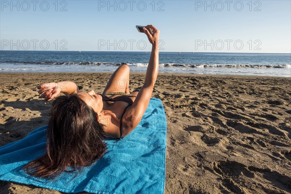 Caucasian woman laying on blanket on beach posing for cell phone selfie