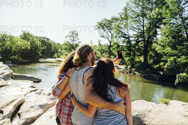 Friends posing for cell phone selfie on rocks near river