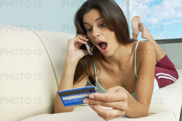 Surprised Hispanic woman laying on sofa with cell phone and credit card