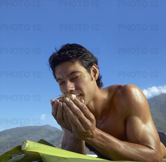 Low angle view of Asian man laying on banana leaves and smelling bowl of botanical medicine