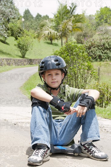 Hispanic boy in pads and helmet sitting on skateboard